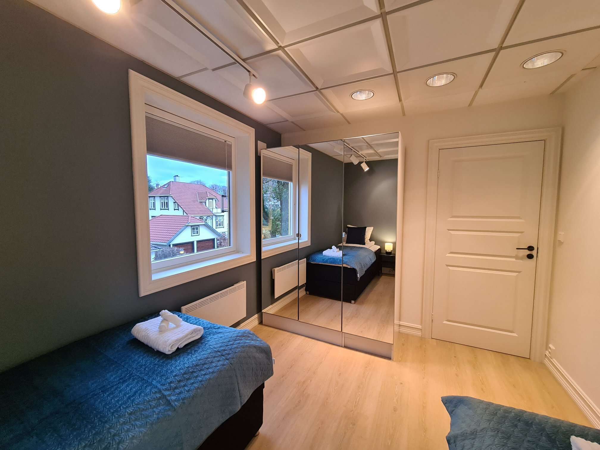 Un dormitorio moderno con un ventanal grande, una cama, una mesita de noche y un espejo que refleja la habitación.