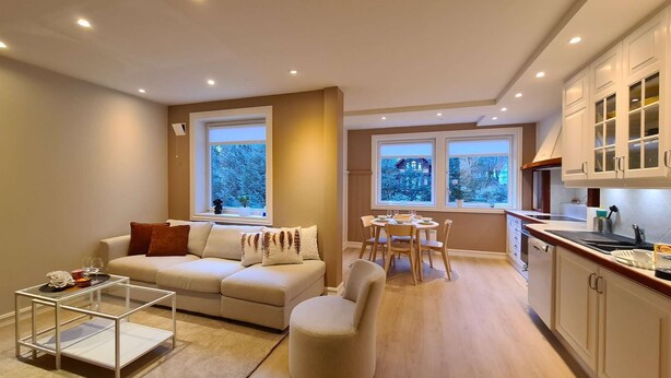Un salon moderne avec un canapé, un pouf et une table basse en verre. On y trouve également un coin repas avec une table et des chaises. La cuisine est dotée de meubles blancs et d’une fenêtre.