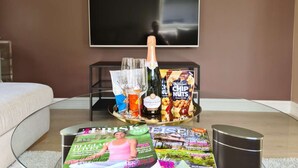 A living room with a flat-screen TV mounted on the wall, a glass coffee table with magazines, a bottle of champagne, and a tray with snacks and glasses. - Bright And Modern - 2-bedroom, Central, Free Parking (Stavanger)