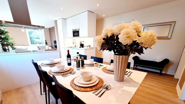 A modern dining area with a white table set for four, surrounded by black chairs. A vase with white flowers is on the table, and there is a bench in the background. - Bright And Modern - 2-bedroom, Central, Free Parking (Stavanger)