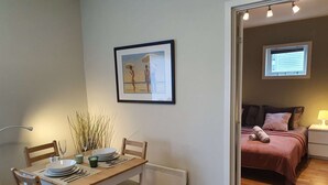 Een eetgedeelte met een tafel gedekt voor twee personen, een ingelijst beeld aan de muur, en een bed met een roze sprei in de aangrenzende kamer.