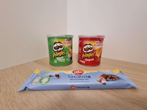 Two Pringles cans, one green and one red, and a blue packet of Firklöver Melsjokolade on a wooden surface. - Room-deluxe Room 4 Rent, King-size Bed, (Stavanger)