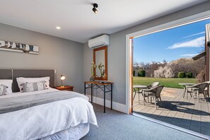 A bedroom with a large bed, bedside table, lamp, and a view of a patio with outdoor furniture. - Manata Homestead and Lodge (Otago)