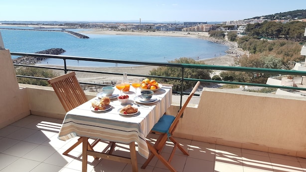 Une table de balcon avec des éléments pour le petit-déjeuner, donnant sur une zone côtière avec une plage et des bâtiments.