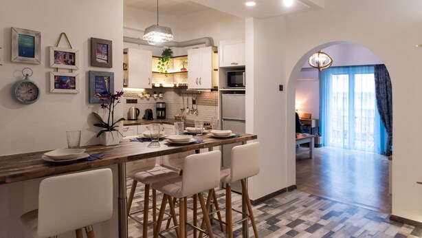 Una cocina moderna con armarios blancos, una isla de madera con taburetes y una zona de comedor con una cortina azul.