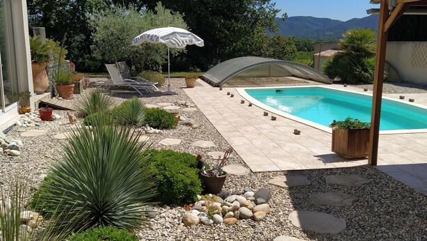 Zona de piscina con vista despejada, rodeada por un sendero de piedrecitas, plantas en macetas y un paraguas blanco.