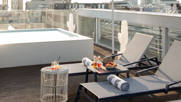Eine Dachterrasse mit blauen Sonnenschirmen, Liegestühlen, einem kleinen Tisch mit Getränken und Blick auf die Stadtlandschaft.