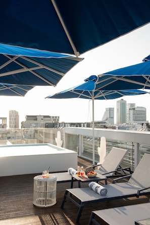 Une terrasse sur le toit, agrémentée de parasols bleus, de chaises longues, d’une petite table avec des boissons, et offrant une vue panoramique sur la ville.