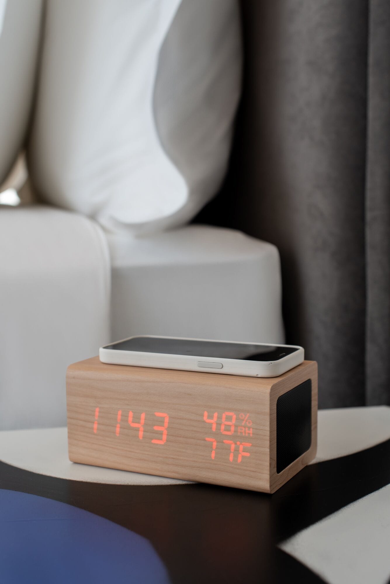 Un comodino in legno con una sveglia digitale che segna le 11:43, un telefono in carica e uno smartphone sopra.