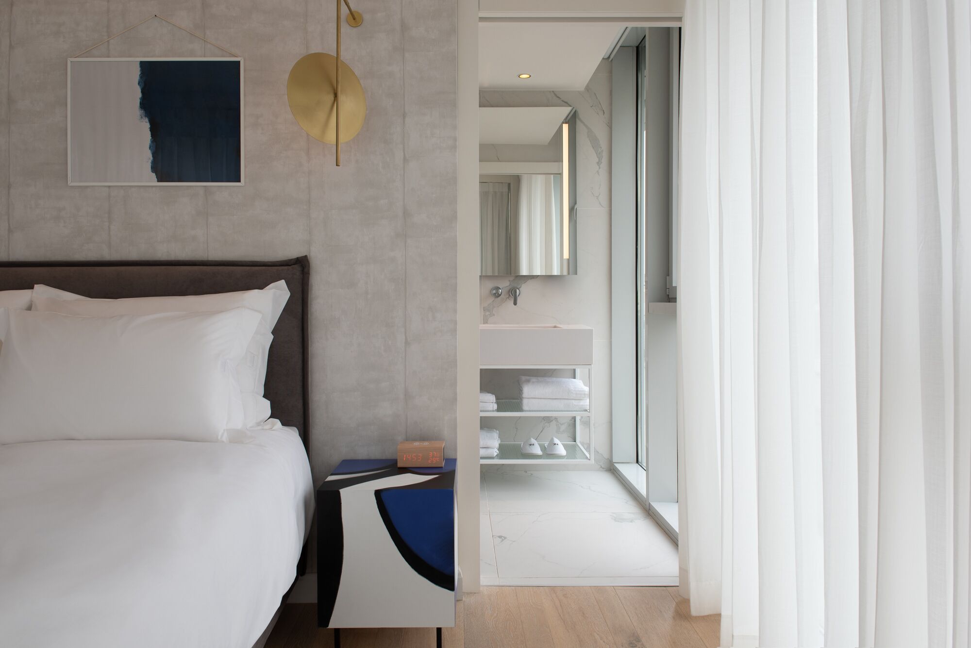 Una habitación de hotel moderna con cama, mesita de noche, espejo, baño con lavabo y toallero, y vistas al exterior.