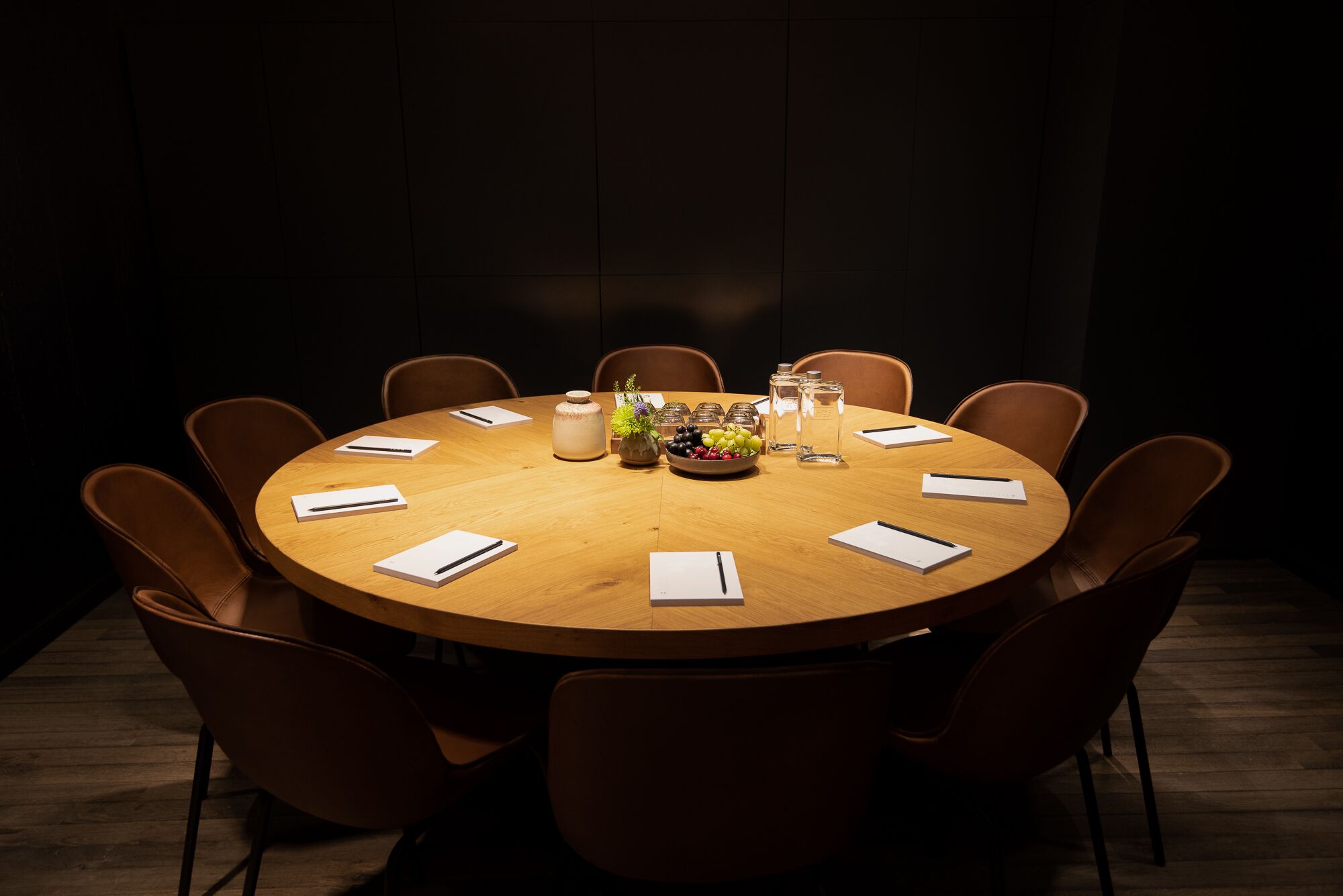 Une table de conférence ronde en bois, entourée de huit chaises marron, un vase rempli de fleurs et un saladier de fruits.