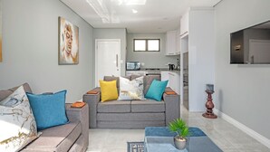 A modern living room with a grey sofa, blue and yellow cushions, a coffee table with a plant, and a kitchen in the background. - Sea De La Vie 25 (La Mercy)