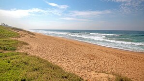A sandy beach with gentle waves, grassy dunes, and a clear sky. - Sea De La Vie 25 (La Mercy)
