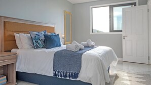 A neatly made bed with white and blue bedding, a wooden nightstand, and a large window with a view of the outdoors. - Sea De La Vie 25 (La Mercy)
