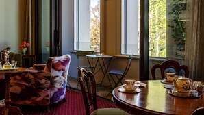 A dining area with a round table set for tea, a chair with a patterned cushion, and a view of trees through large windows. - Gaynor Clayton Apartment (Dunedin)
