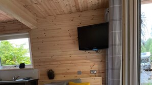 A cozy cabin interior with a wooden table, a sofa with cushions, a fluffy white stool, and a television mounted on the wall.