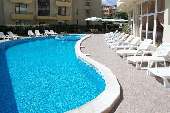 A swimming pool with white lounge chairs and umbrellas, surrounded by a building with balconies and windows.