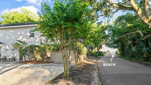 Una calle residencial con una casa, un árbol y una acera que lleva a la playa.