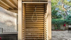 Una ducha exterior de madera con manguera, rodeada por un muro de piedra y un suelo de madera.