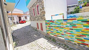 A narrow cobblestone street with a stone building featuring pink-framed windows and a colorful wall. - Otel Meryem Ana Bozcaada (Bozcaada)