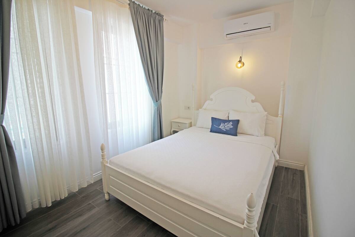 A bedroom with a bed, curtains, a bedside table, and an air conditioning unit.