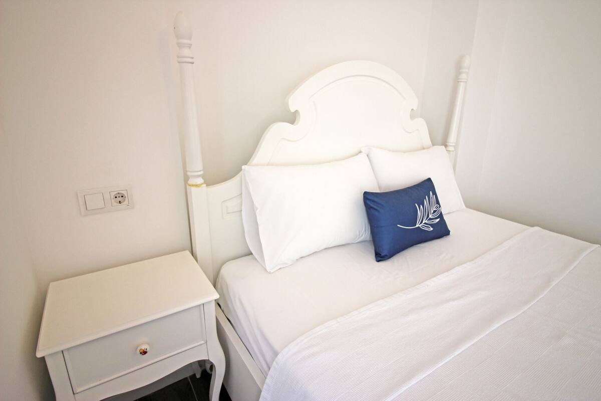A neatly made bed with white bedding and a blue pillow, a white nightstand with a drawer, and a white wall with a power outlet.