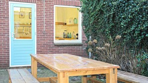 A wooden table and bench set against a brick wall with a window and a green hedge.