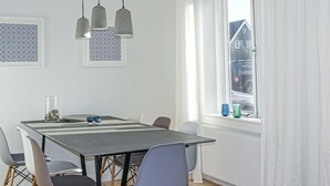A modern dining area with a long table, grey chairs, pendant lights, and framed artwork on the wall.