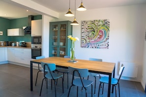 A modern kitchen with teal cabinetry, a wooden dining table, and blue chairs. There is a colorful abstract painting on the wall and a vase with flowers on the table.