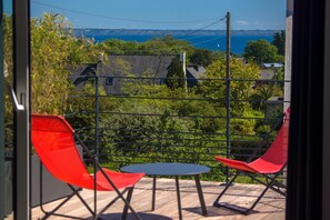 A balcony with two red chairs and a small table, overlooking a coastal landscape with houses and trees.