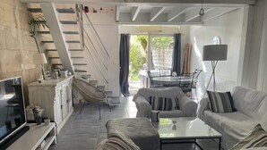 Een moderne woonkamer met een bank, fauteuil en salontafel. Er is een trap naar een hoger gelegen verdieping, een eethoek en een tv op een standaard.