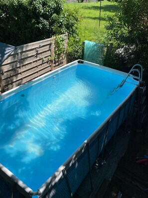 A rectangular swimming pool with a ladder on one side, surrounded by a wooden fence and greenery.