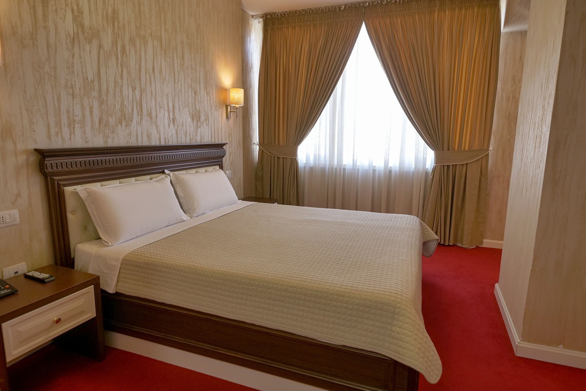 Une chambre d’hôtel avec un grand lit, une table de chevet avec une lampe et une vue sur l’extérieur à travers des rideaux transparents.