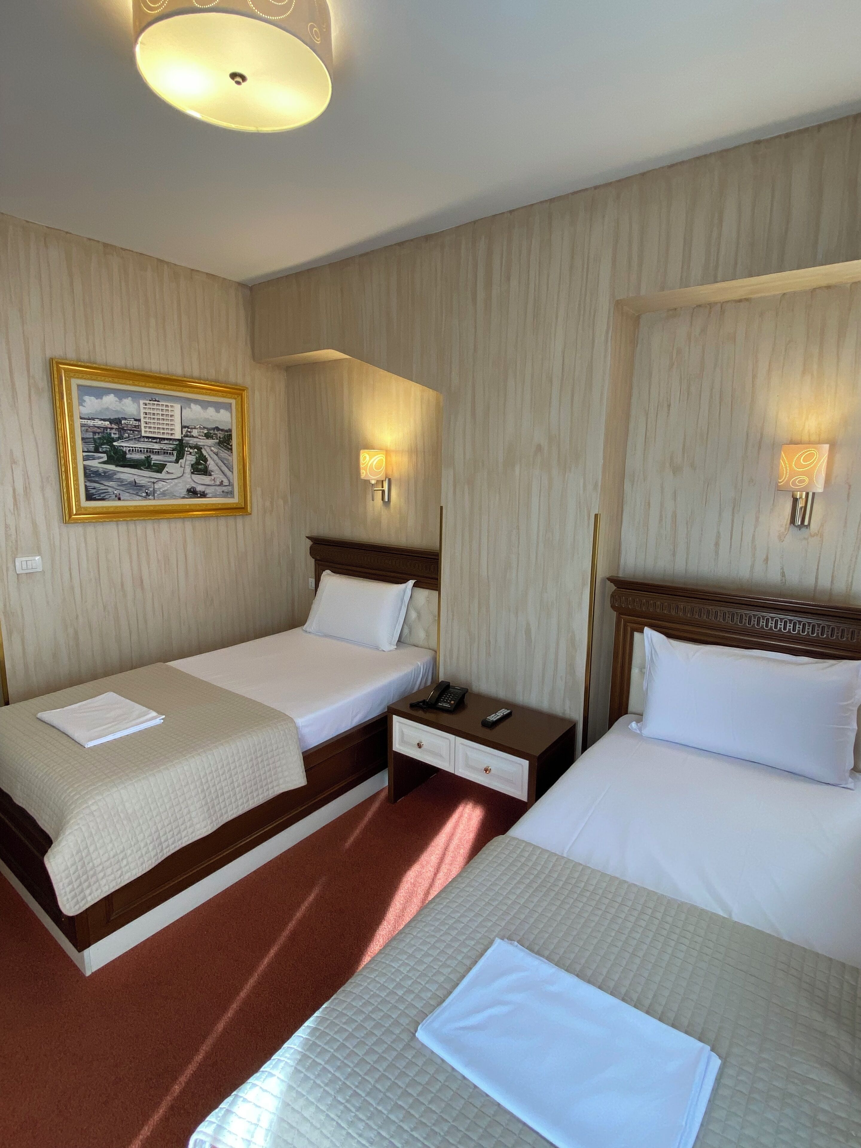 A hotel room with two beds, a nightstand with a phone and remote, a framed picture on the wall, and a ceiling light.