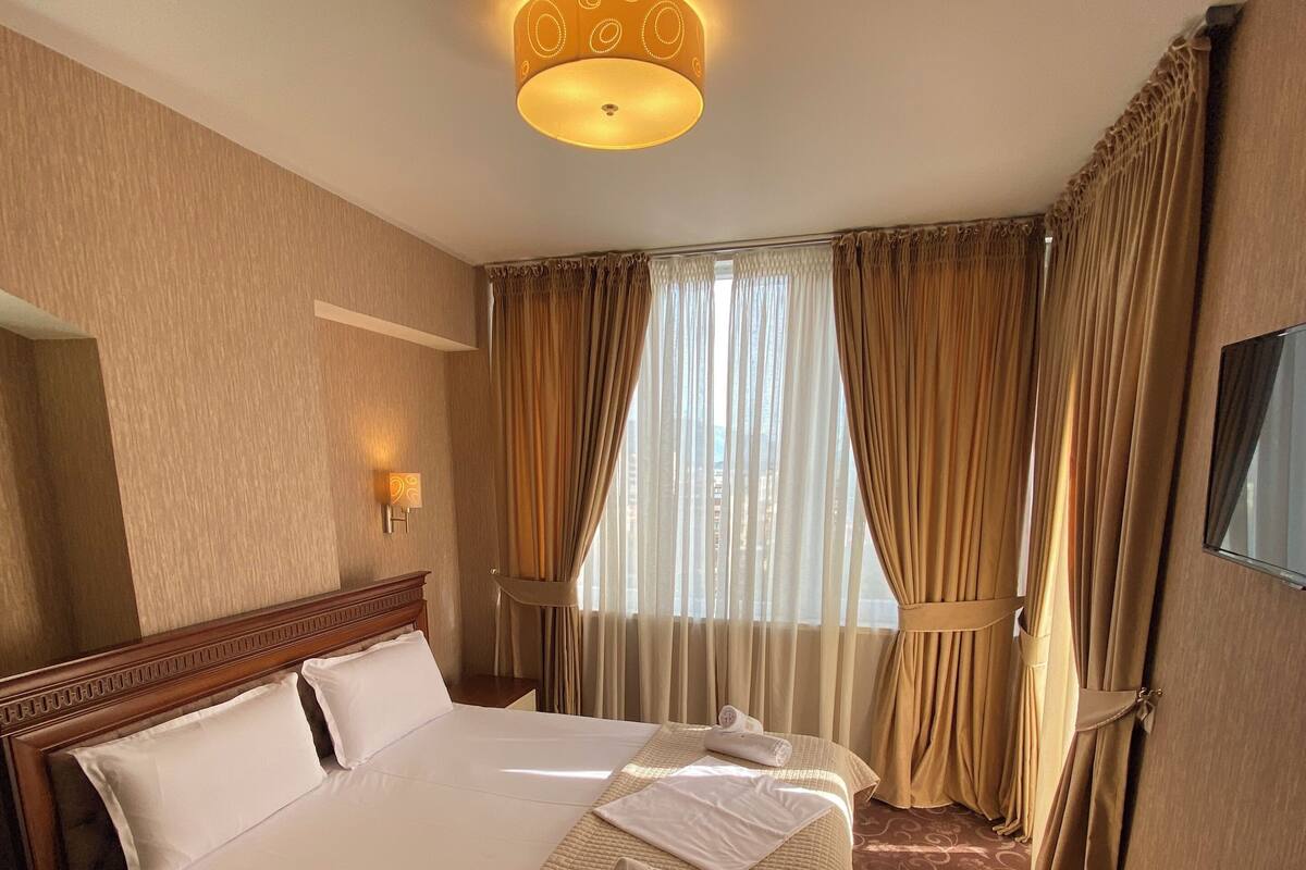 Ein Hotelzimmer mit einem großen Bett, einem Nachttisch, einer Lampe und einem Fenster mit Vorhängen.