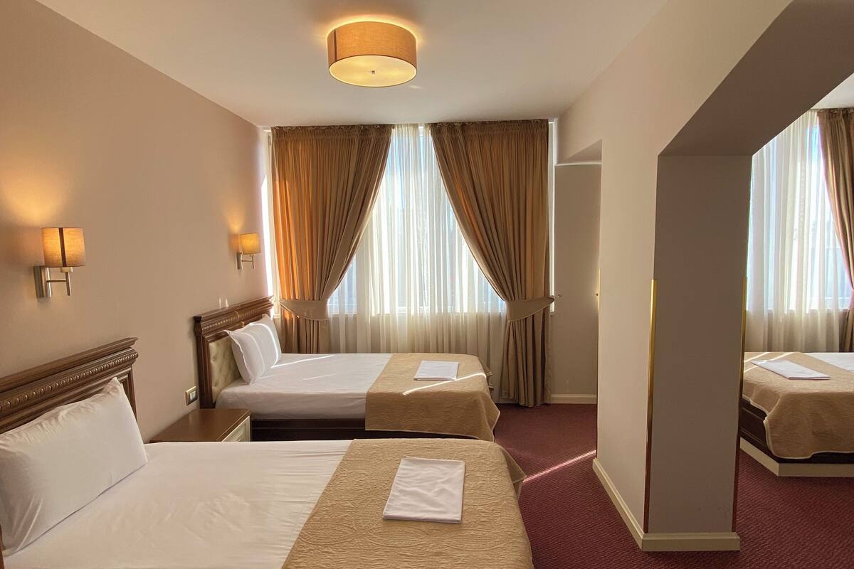 Ein Hotelzimmer mit zwei Betten, einem großen Fenster mit Vorhängen, einer Deckenlampe und einem Teppichboden.