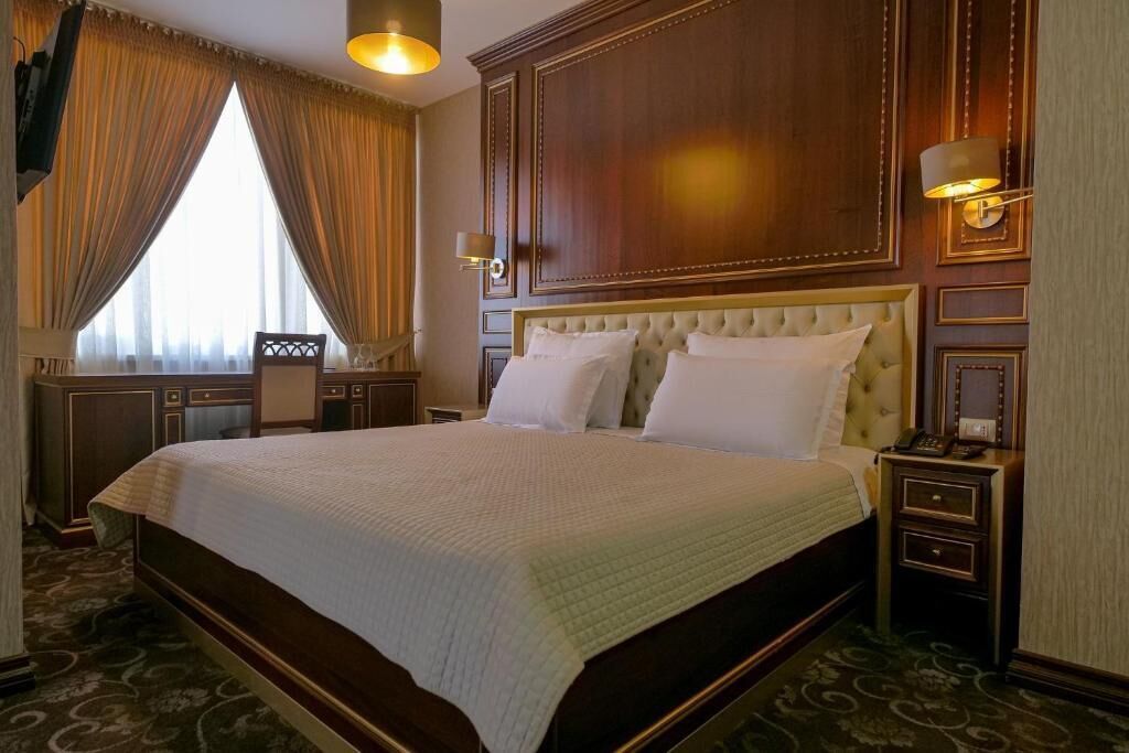 Una cama grande con cabecera capitonada, una mesita de noche con lámpara y vistas al exterior a través de una ventana con cortinas.