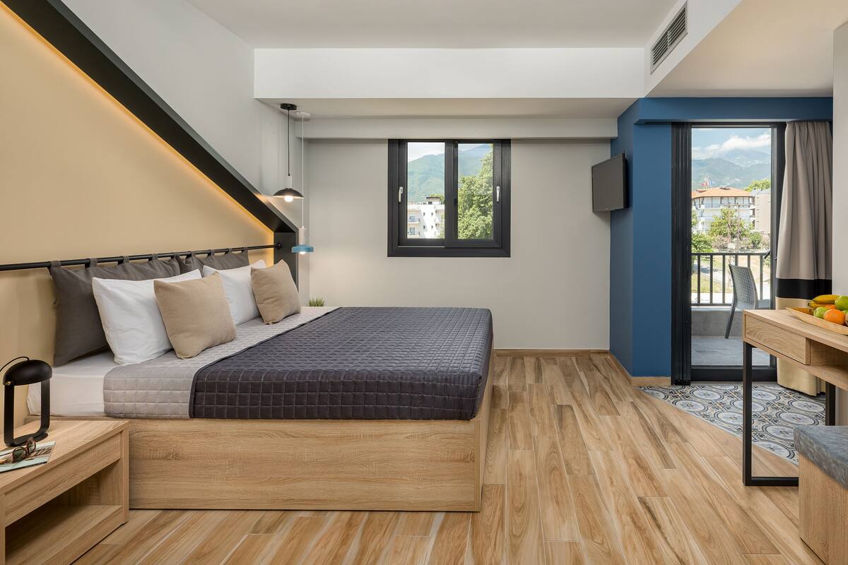 A modern hotel room with a bed, bedside table, and a desk with a chair. There is a window with a view of buildings and mountains.