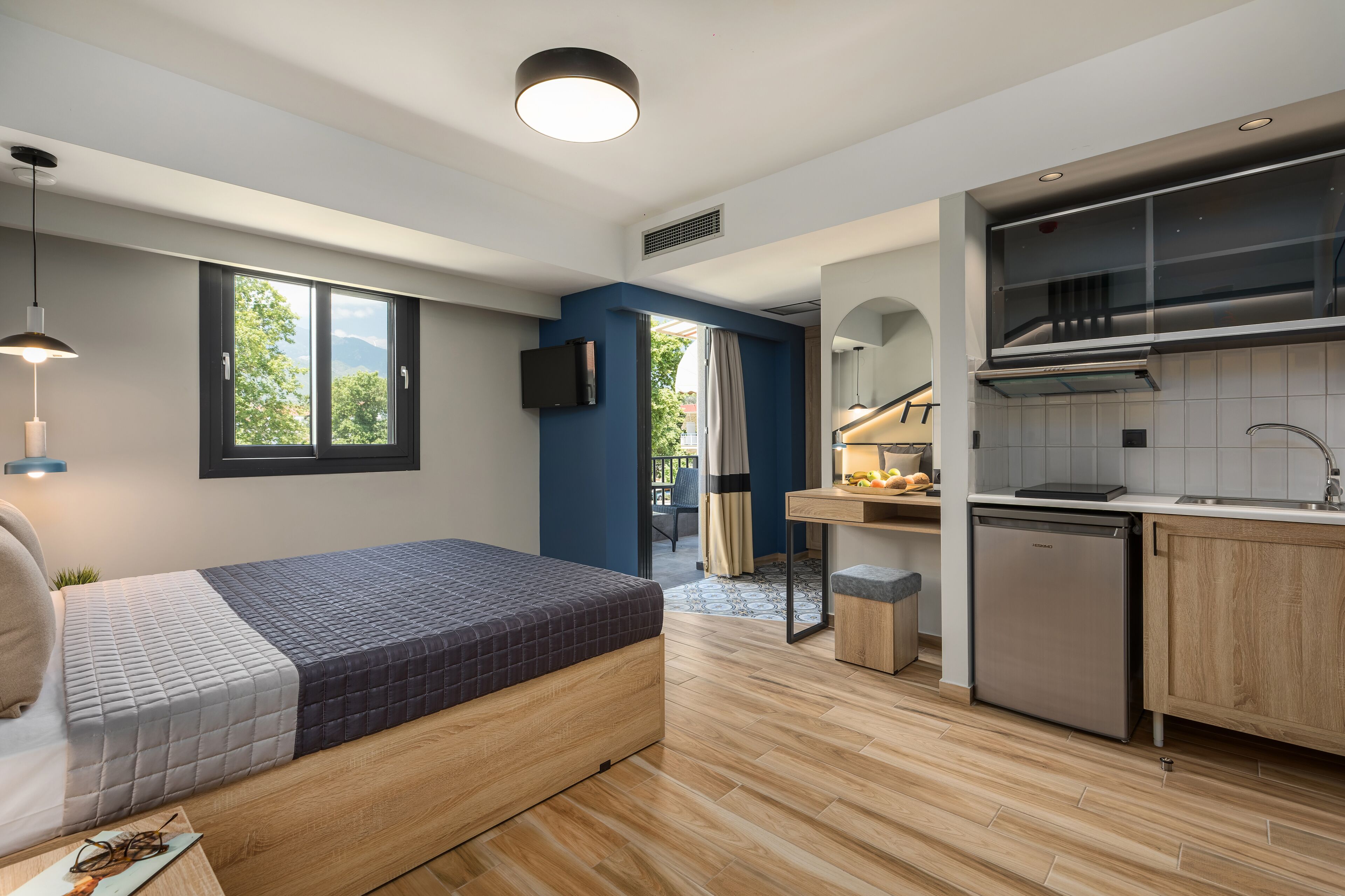A modern, compact hotel room with a bed, kitchenette, and a view of the outdoors.