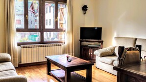 Una sala de estar con sofá, mesa de centro, televisor sobre un mueble bajo y ventana con vista a edificios.
