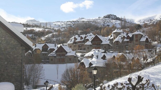 Ein verschneites Dorf mit traditionellen Holzhäusern, einem Steinhaus und einer Straßenlaterne.