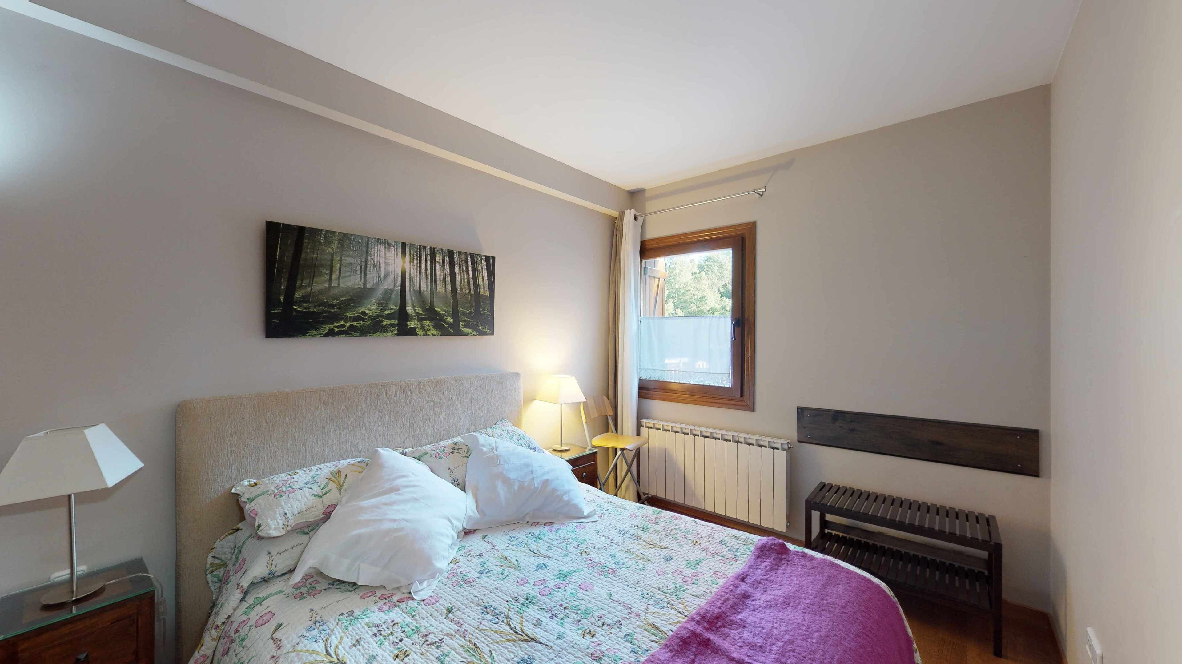 Une chambre à coucher avec un lit, une table de chevet, une lampe, un radiateur, une fenêtre et un tableau accroché au mur.