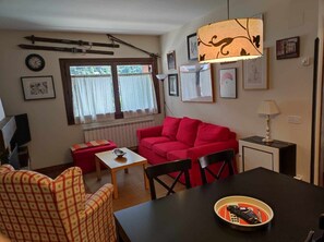 A living room with a red sofa, a small table, and a coffee table. There is a window with curtains, a radiator, and a lamp on a side table.