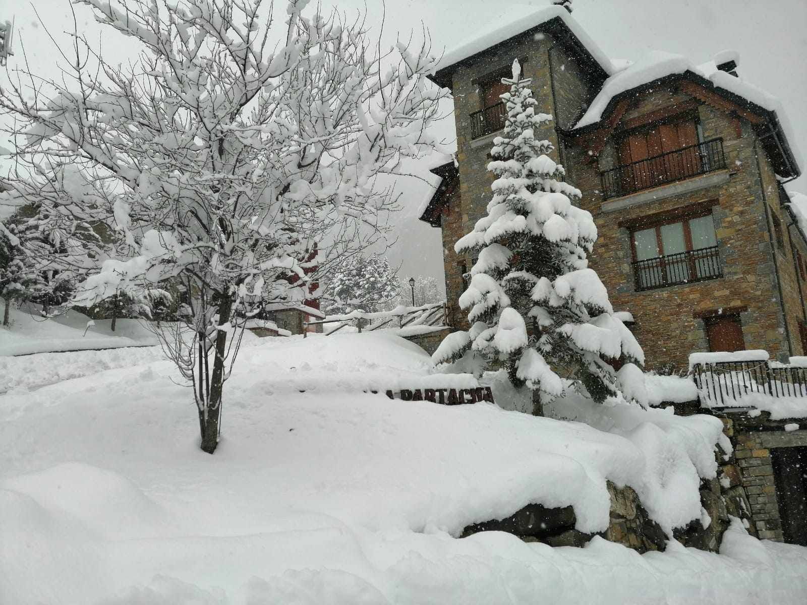 A snow-covered building with a stone facade, a balcony, and a snow-covered tree in front.