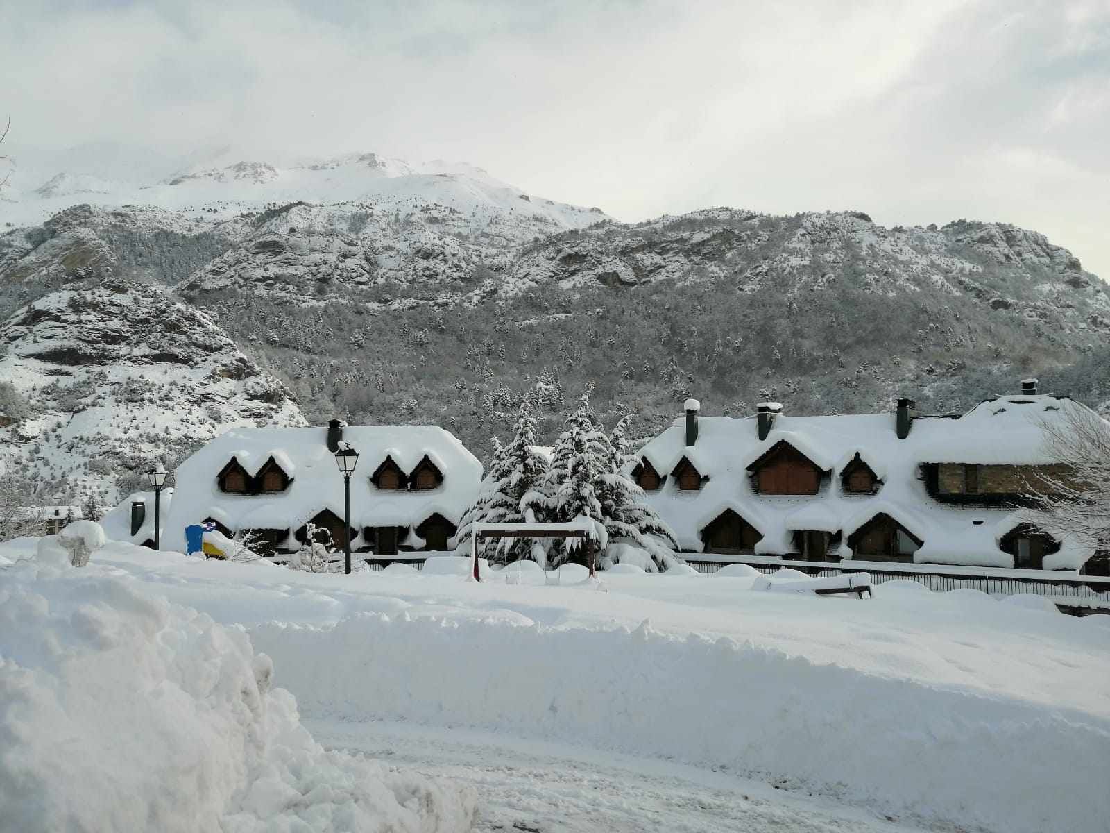 Un albergue cubierto de nieve con varias cabañas de madera, un árbol central y un paisaje montañoso de fondo.