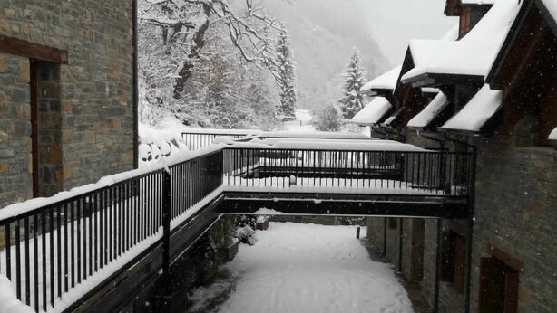 Ein verschneiter Innenhof mit einer Holzbrücke, Ziegelgebäuden und mit Schnee beladenen Bäumen.