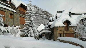 Ein mit Schnee bedecktes Gebäude mit Steinmauern und hölzernen Balkonen, umgeben von schneebedeckten Bäumen und einem Berg im Hintergrund.