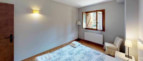 Un dormitorio con cama, armario de madera, ventana con vistas, radiador y mesita de noche con lámpara.