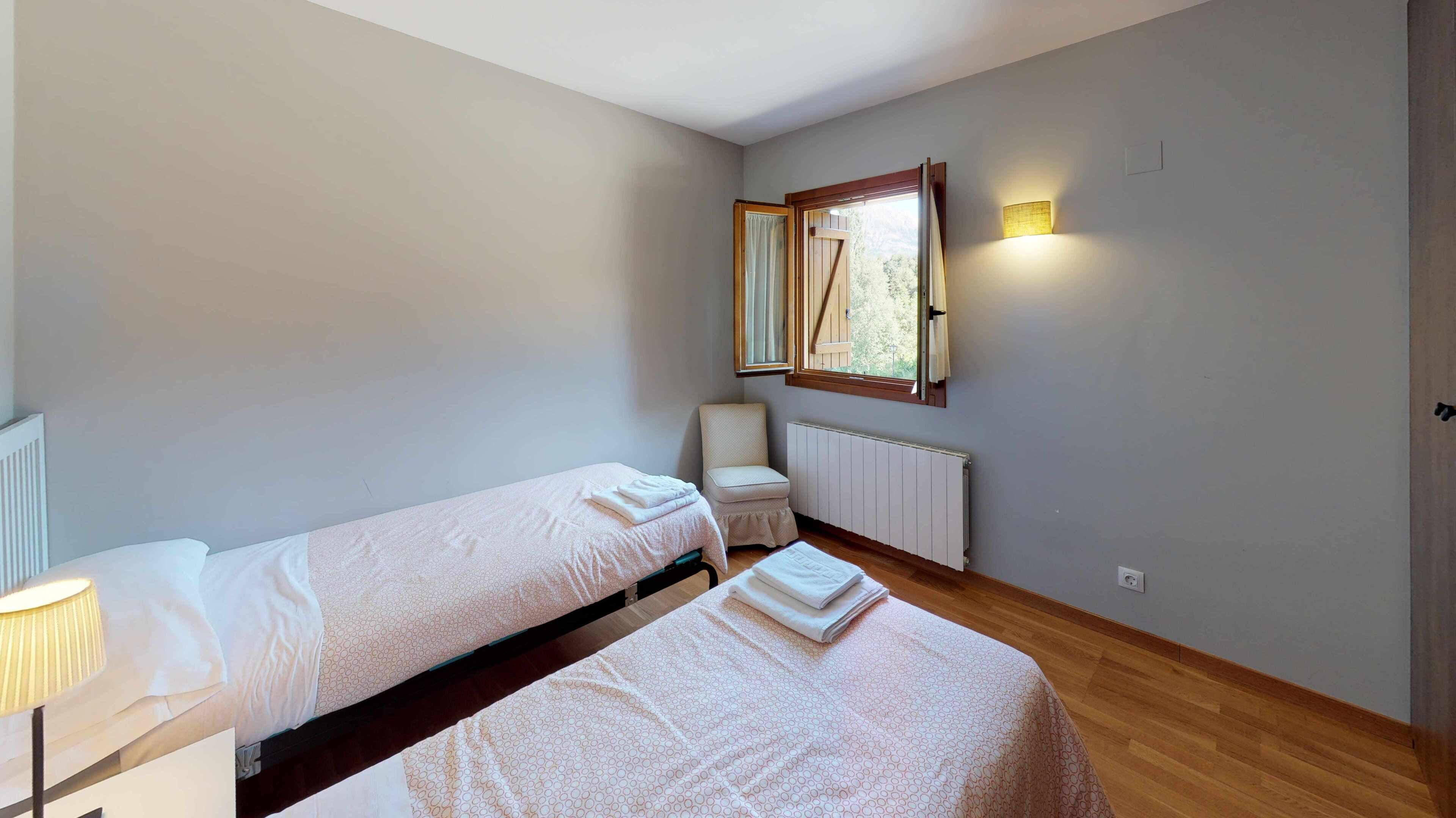 Une petite pièce bien éclairée, avec deux lits simples, une chaise, un radiateur et une fenêtre avec des rideaux.