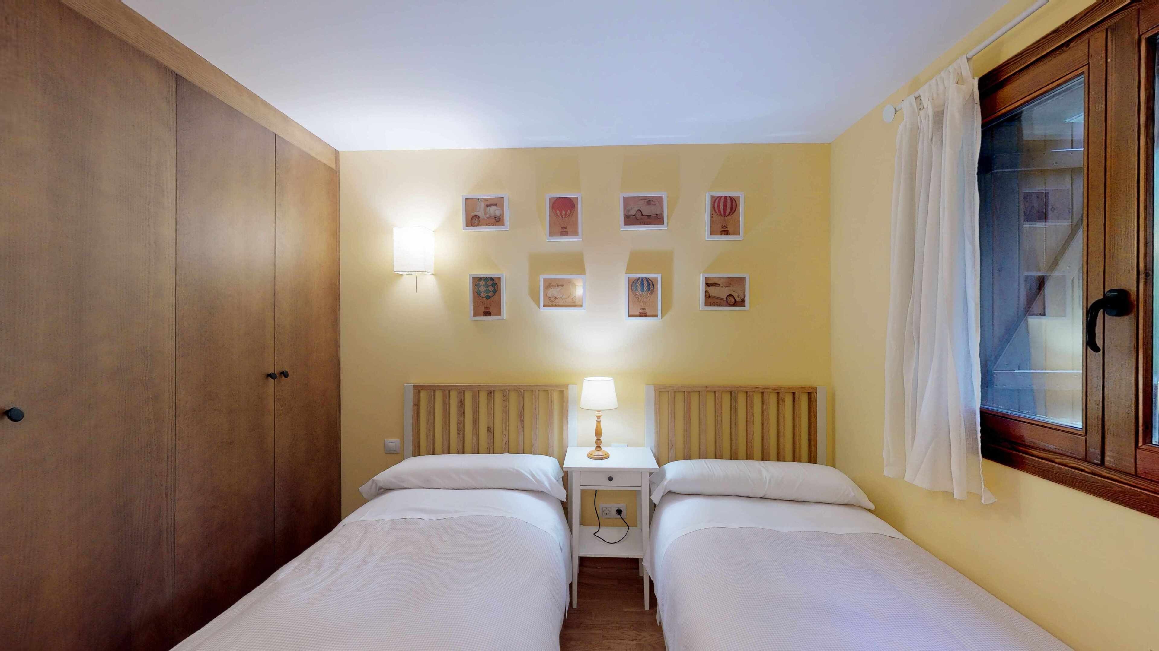 Een kamer met twee bedden, een houten garderobe, een tafeltje met een lamp en een raam met gordijnen.